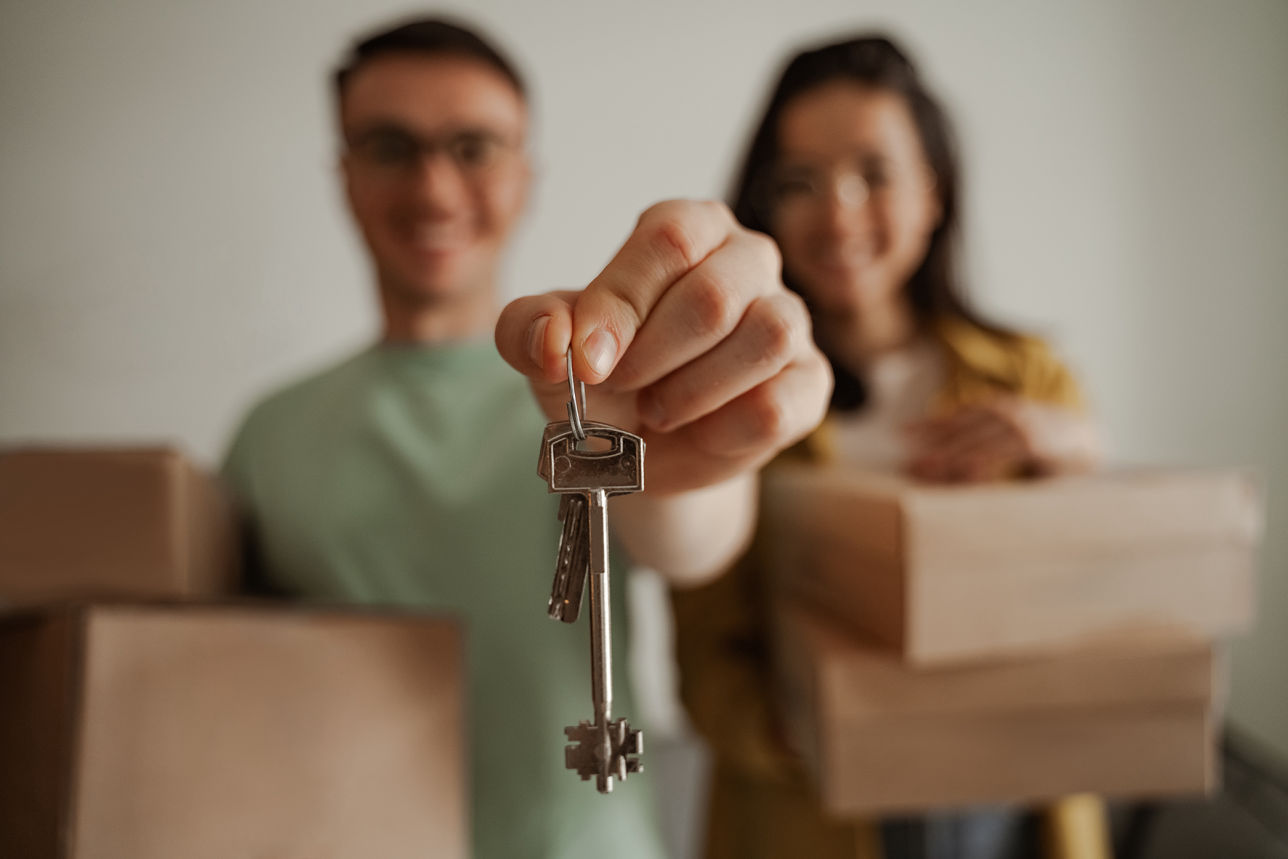 Pareja joven entregando las llaves de su nueva casa con cajas de mudanza al fondo